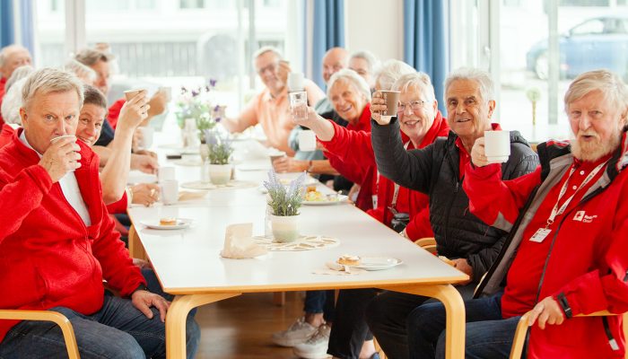 En gruppe personer som sitter ved et bord. Alle reiser kaffekoppene sine og smiler.
