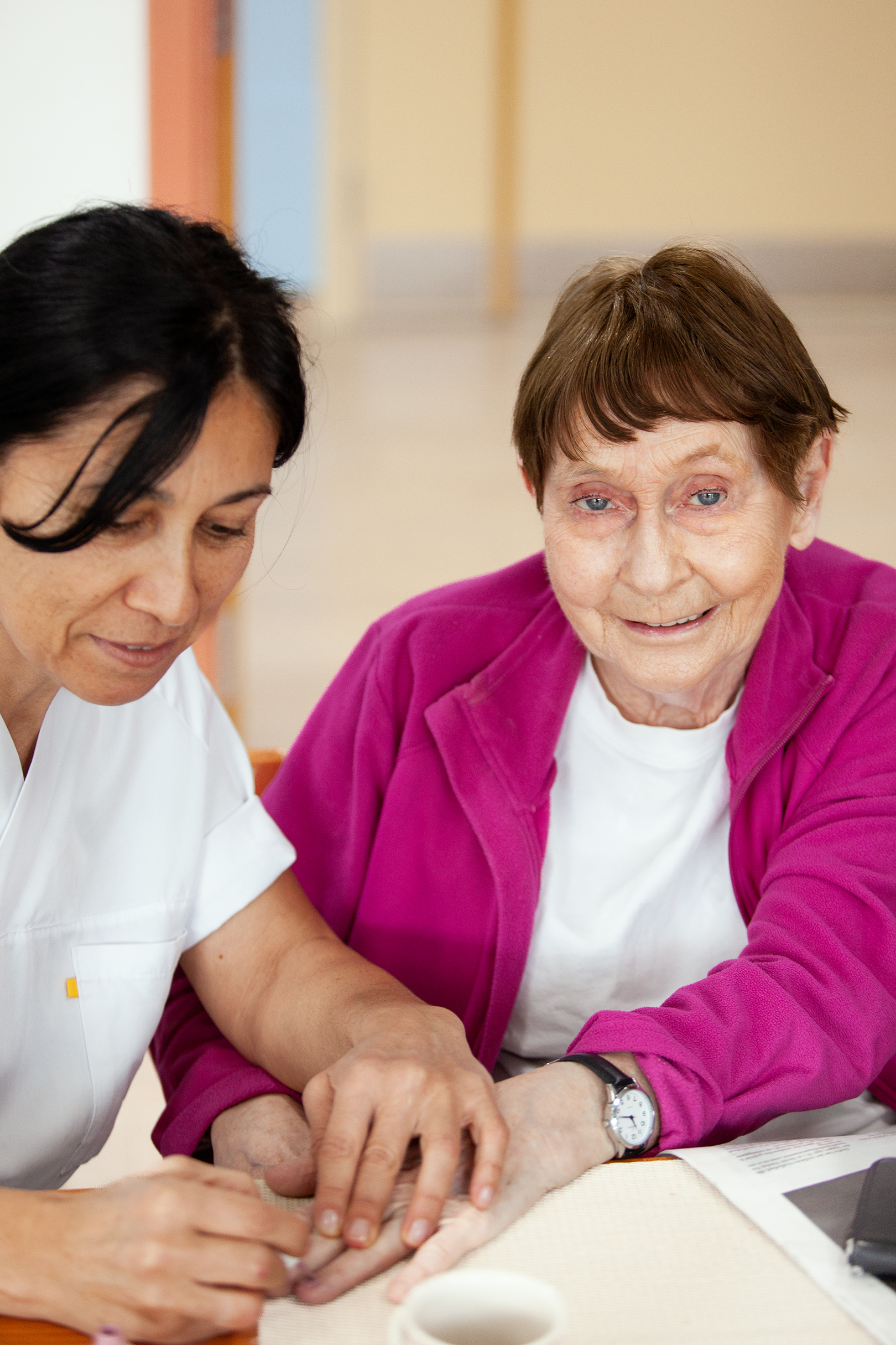 To personer som sitter ved et bord. Den ene personen hjelper den andre personen med å få rengjort neglene sine.